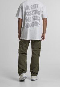 Camiseta gris con gran texto en la espalda, combinada con pantalones cargo verde oliva y zapatillas blancas con detalles amarillos. Conjunto de ropa casual.