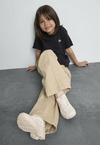 Svart T-skjorte, beige cordfløyelsbukser og hvite sneakers. Barn som sitter mot en vegg, bena strukket ut, med et lite smil.
