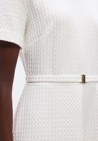 Gros plan d'une personne portant une robe blanche texturée à manches courtes et une fine ceinture blanche avec une petite boucle dorée à la taille.