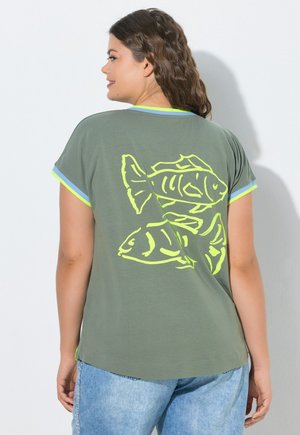 Femme portant un t-shirt vert à manches courtes avec un graphisme de poisson jaune néon dans le dos et un jean bleu clair, debout contre un mur blanc.