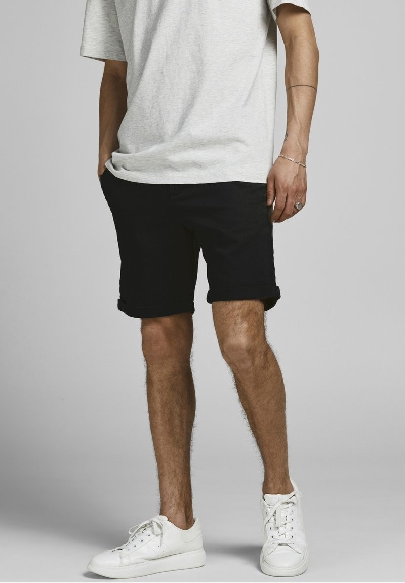 Homme portant un t-shirt gris clair, un short noir retroussé, des baskets blanches, debout avec une main dans la poche devant un fond uni.