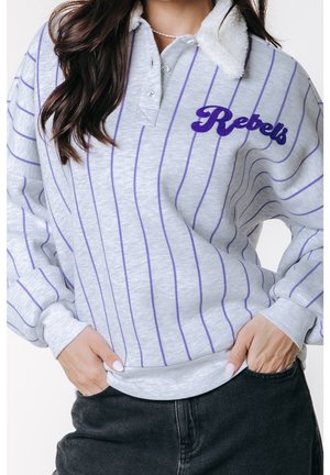 Kvinde iført en lys grå pullover med lilla lodrette striber, "Rebels" broderet på brystet, hvid sherpa-krave og sorte jeans, hænderne i lommerne.