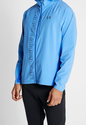 Mann trägt hellblaue Under Armour Jacke mit Reißverschluss und vertikalem Logo auf der linken Brustseite und schwarze Hose vor neutralem Hintergrund.