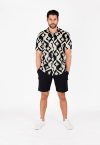 Schwarz-weiß gemustertes Kurzarmhemd mit geometrischen Designs, kombiniert mit schwarzen Shorts und weißen Sneakers. Modell steht mit Händen in den Taschen.