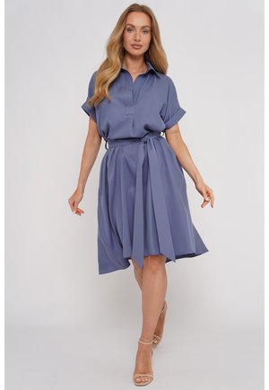 Kobieta z długimi blond włosami, ubrana w niebieską sukienkę z krótkim rękawem i paskiem oraz beżowe sandały na wysokim obcasie, stoi na jednolitym tle.