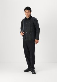 Black, textured coat with a button-up front, and two front pockets. Worn over dark pants, paired with black shoes, and a shoulder strap.