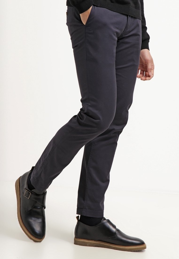 Modèle avec jambes portant un pantalon noir ajusté et des chaussures en cuir noires aux semelles marron, une main dans la poche, pose en marchant sur fond blanc.