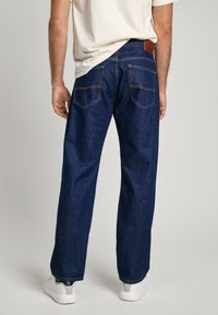 Jeans de denim azul oscuro con un corte relajado, que cuentan con bolsillos traseros con detalles de costura y un parche de cuero marrón en la cintura.