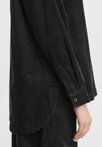 Camisa de pana negra con puños abotonados y dobladillo redondeado, que destaca por su textura acanalada y su acabado suave.
