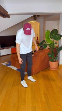 Balts T-krekls, melnas cargo bikses un baltas tenisa kurpes ar dzelteniem akcentiem. Valdzina burgundijas krāsas cepure. Telpas iekārtojums ar koka grīdu un augu.