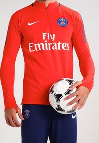 Maillot à manches longues rouge Nike avec col montant, arborant le logo "Fly Emirates" et le badge du PSG. Un pantalon bleu et un ballon de football noir et blanc sont tenus.