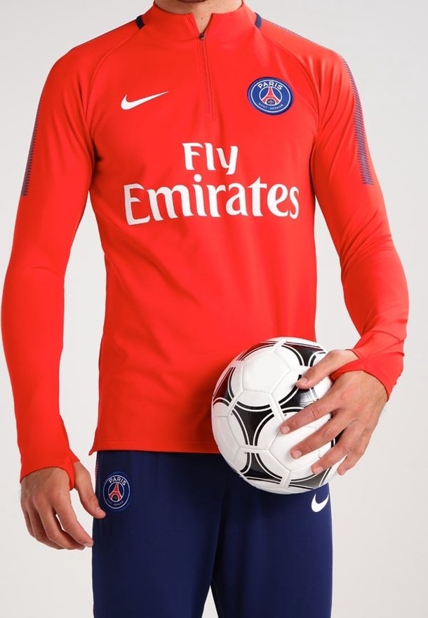 Maillot à manches longues rouge Nike avec col montant, arborant le logo "Fly Emirates" et le badge du PSG. Un pantalon bleu et un ballon de football noir et blanc sont tenus.