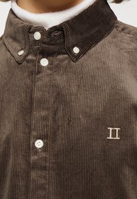 Camisa de pana marrón con cuello abotonado, que cuenta con botones blancos y un sutil bordado "II" en la parte izquierda del pecho.