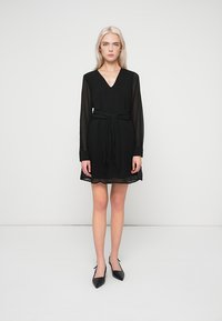 Μαύρο φόρεμα με μακριά διάφανα μανίκια και V-λαιμόκοψη. Διαθέτει ζώνη που δένει στη μέση, μήκος πάνω από το γόνατο και ελαφρώς φουσκωτό, υφαντό ύφασμα.