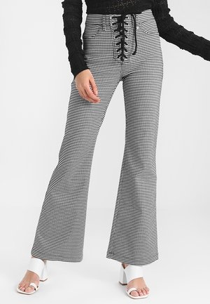 Schwarz-weiß kariert Hose mit hohem Bund und vorderer Schnürung, kombiniert mit einem schwarzen, strukturierten Langarmoberteil und weißen Absatzsandalen.