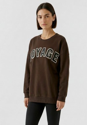 Vrouw met kort bruin haar die een donkerbruine trui draagt met "VOYAGE" in grote letters op de borst gedrukt en zwarte broek.