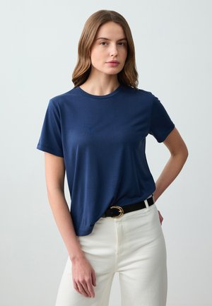 T-shirt bleu marine avec un col rond et des manches courtes, en tissu lisse. Associé à un pantalon blanc et à une ceinture noire avec une boucle en or.