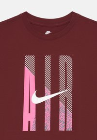 Bordo majica s kratkim rukavima koja sadrži boldirani natpis "AIR" u ružičastoj i bijeloj boji s prugastim i uzorkovanim detaljima; Nike logo integriran u dizajn.