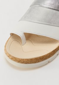 Bela in srebrna drsna sandal, ki ima teksturirano podplato, tkane plute podrobnosti na podplatu in odprto obliko prstov.