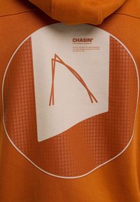 Oranje hoodie met een cirkelvormige graphic op een gestructureerde, oranje achtergrond en een witte panel met gestileerde lijnen en tekst.