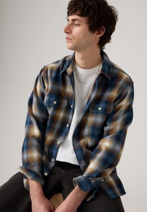 Flannel shirt featuring a blue, brown, and white check pattern, with button front and chest pockets, worn over a white shirt. Black jeans.