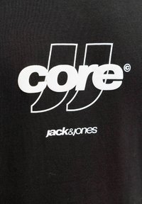 Črna bombažna majica s kratkimi rokavi z velikim belim stiliziranim napisom "core" z dvema ukrivljenima poudarkoma in manjšim napisom "jack & jones" pod njim.