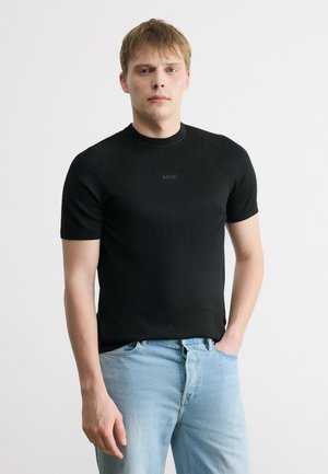 Giovane uomo con maglione nero a maniche corte con logo "BOSS" e jeans azzurri, in piedi con una mano in tasca, sfondo neutro.