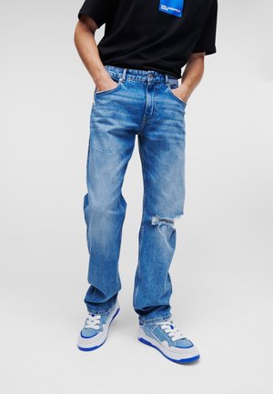 Personne portant un jean bleu clair déchiré, des baskets bleues et blanches, et un t-shirt noir avec un petit logo bleu sur la poitrine.