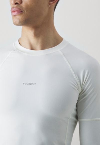 Fehér, hosszú ujjú sportpóló, rugalmas anyagból készült, lapos varrásokkal és egy diszkrét "soulland" logóval az elején.