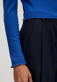 Nahaufnahme einer Person, die ein strukturiertes, blaues Langarmoberteil und schwarze hochgeschnittene Hosen trägt, mit Details zu Arm und Hand.