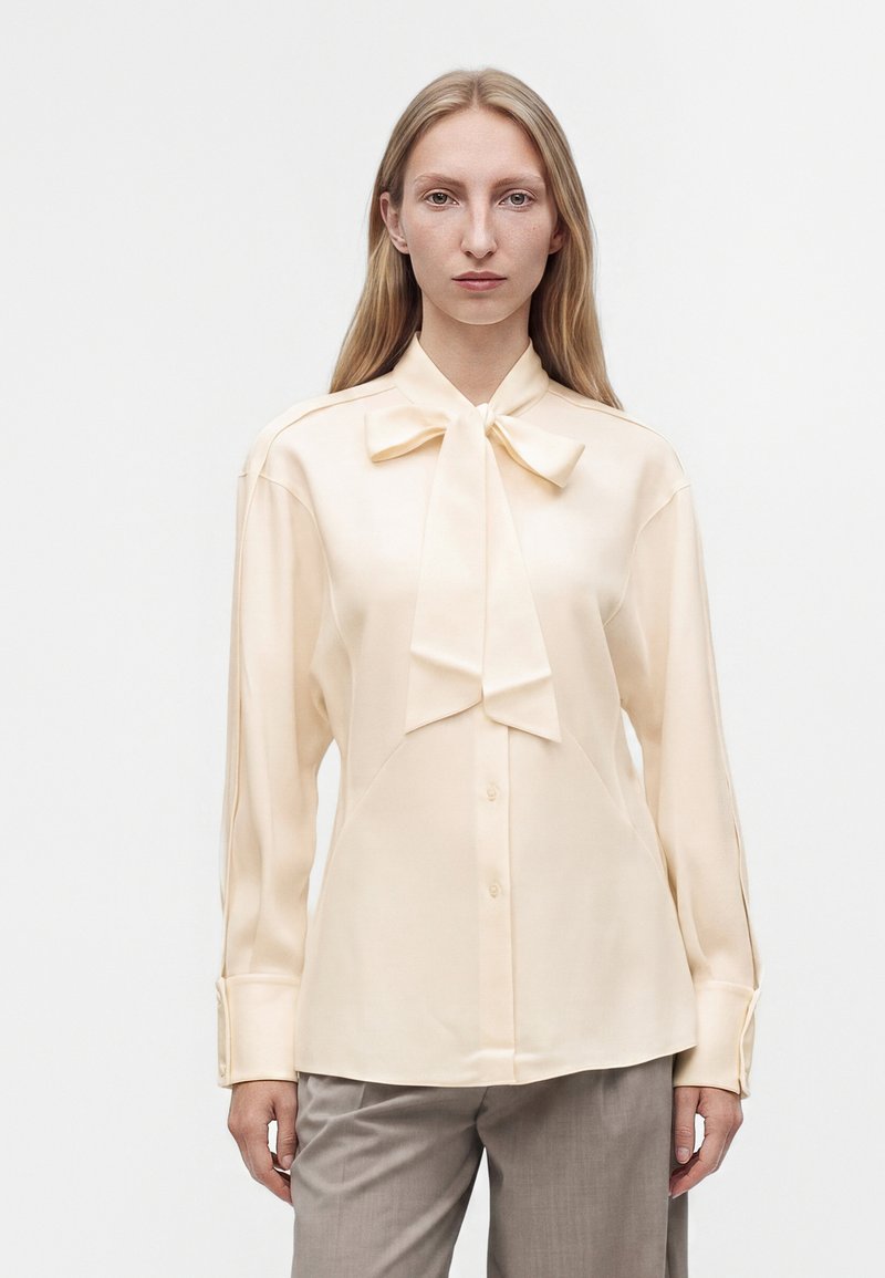 Femme aux longs cheveux blonds portant un chemisier crème avec un grand nœud au col et un pantalon gris, debout sur fond blanc.