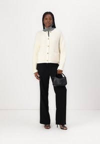Cardigan blanc en peluche avec des boutons dorés et des poches, associé à un pantalon noir et des talons avec des brides. Tenant un petit sac à main noir.