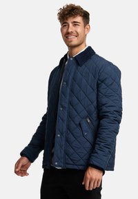 Quilted navy jacket with corduroy collar and zippered side pocket. Features snap buttons and textured fabric, offering a structured design.