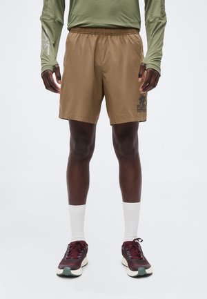 Mężczyzna noszący jasnobrązowe szorty z logo "Wild By Nature" na lewej nodze, zieloną bluzę z długim rękawem, białe skarpetki i ciemnoczerwone buty do biegania.