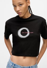Melnā īsā t-krekls, kas izgatavots no kokvilnas, ar apļveida baltu grafiku un sarkanu tekstu "KARL LAGERFELD JEANS" krūšu daļā.