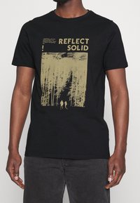Svart bomulls t-shirt med en stor gyllene grafisk tryck som föreställer träd och figurer, med texten "REFLECT SOLID" högst upp.