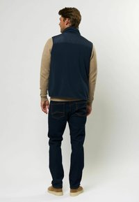 Marinblå fleeceväst med hög krage, beige tröja under, mörka jeans i denim och bruna mockaskor. Enkelt design utan synliga mönster.