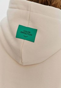 Béžová mikina s texturovaným povrchem, na zadní straně s zelenou nálepkou "COLOR R3VOLUTION". Jednoduchý design a moderní tvar.