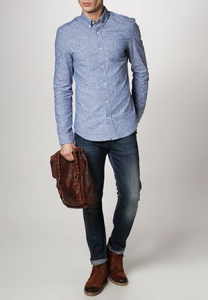 Mężczyzna w jasnoniebieskiej koszuli w drobny wzór zapinanej na guziki, ciemnych dżinsach i brązowych skórzanych butach, trzymający brązową skórzaną torbę z plecionymi uchwytami.