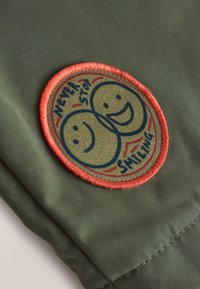 Parche de tela redondo con dos caras sonrientes superpuestas y texto "NEVER STOP SMILING" sobre fondo de tela verde.