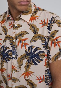 Camicia a maniche corte realizzata in tessuto beige decorato con grandi motivi a foglia verdi e blu navy, arricchita da dettagli floreali arancioni e bottoni bianchi.