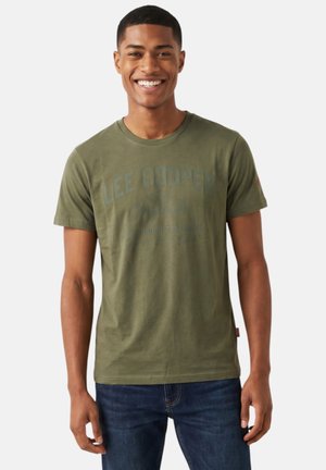 Homme souriant portant un t-shirt vert olive Lee Cooper et un jean bleu foncé, debout devant un fond clair uni.