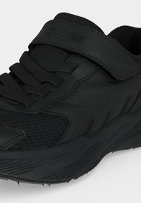 Chaussure de sport noire avec tige en mesh et synthétique, col matelassé, fermeture à lacets à l’avant et bande auto-agrippante sur le dessus.