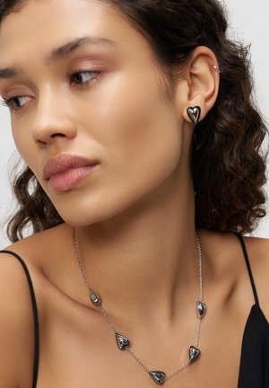 Frau mit lockigem Haar, die schwarze herzförmige Ohrringe und eine passende Halskette an einer Silberkette trägt, gekleidet in einem schwarzen Spaghettiträger-Top.