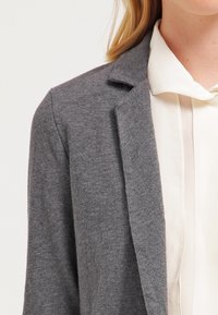 Grå blazer gjord av mjukt tyg med kavajsnibb, enkelknappsstängning och lätt strukturerad yta, lagd över en krämfärgad blus.