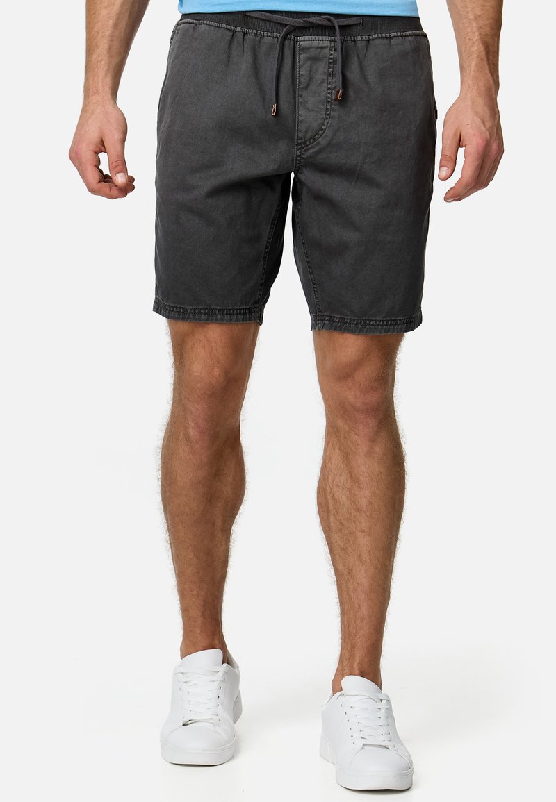 Homme portant un short gris foncé avec cordon de serrage et des baskets blanches, debout devant un fond blanc.