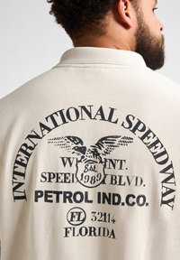 Beige polo shirt met een zwarte grafische print op de achterkant, met de tekst "INTERNATIONAL SPEEDWAY" en een adelaarsmotief met aanvullende tekst.