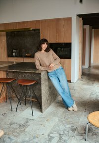 Pull en maille beige avec des boutons, jeans d'audacieuse coupe large bleu clair, et baskets beiges. Plan de travail en pierre, armoires en bois, et carrelage gris visible.