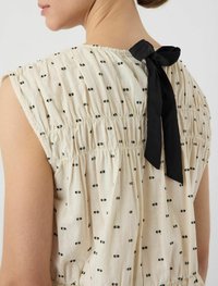 Vestido de tela cremosa con patrones de ojal negros. Cuenta con un escote fruncido y un lazo de cinta negra en la parte posterior. Diseño sin mangas.