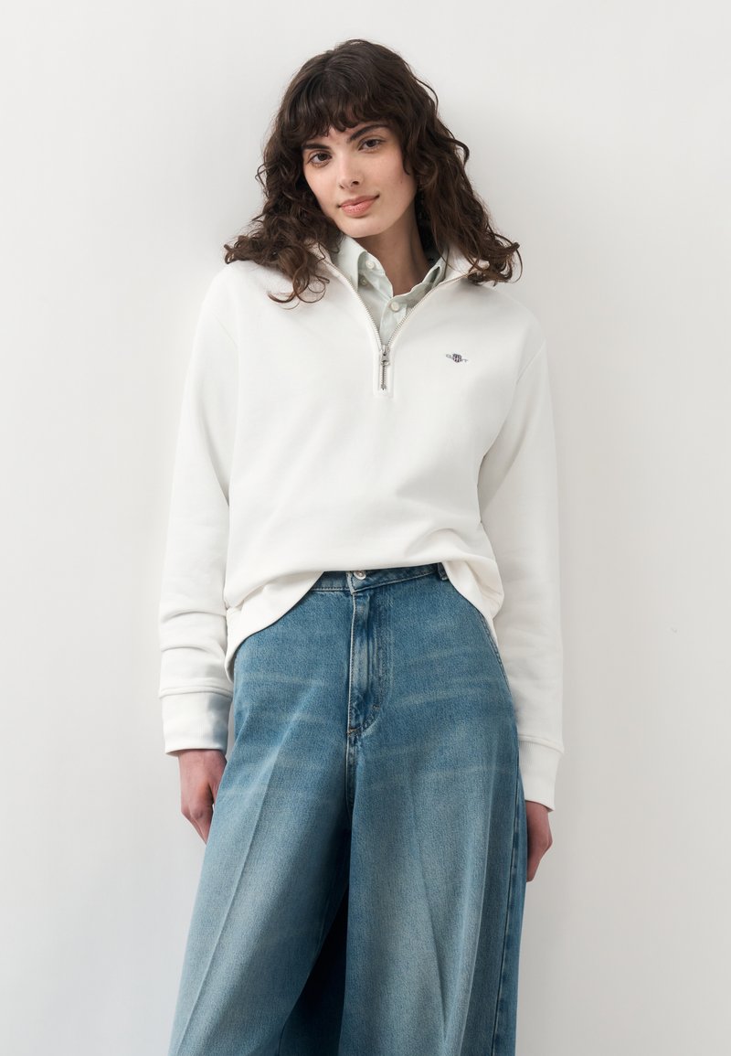 Jeune femme aux cheveux bouclés foncés portant un sweat blanc à zip partiel sur une chemise claire et un jean bleu ample, debout.